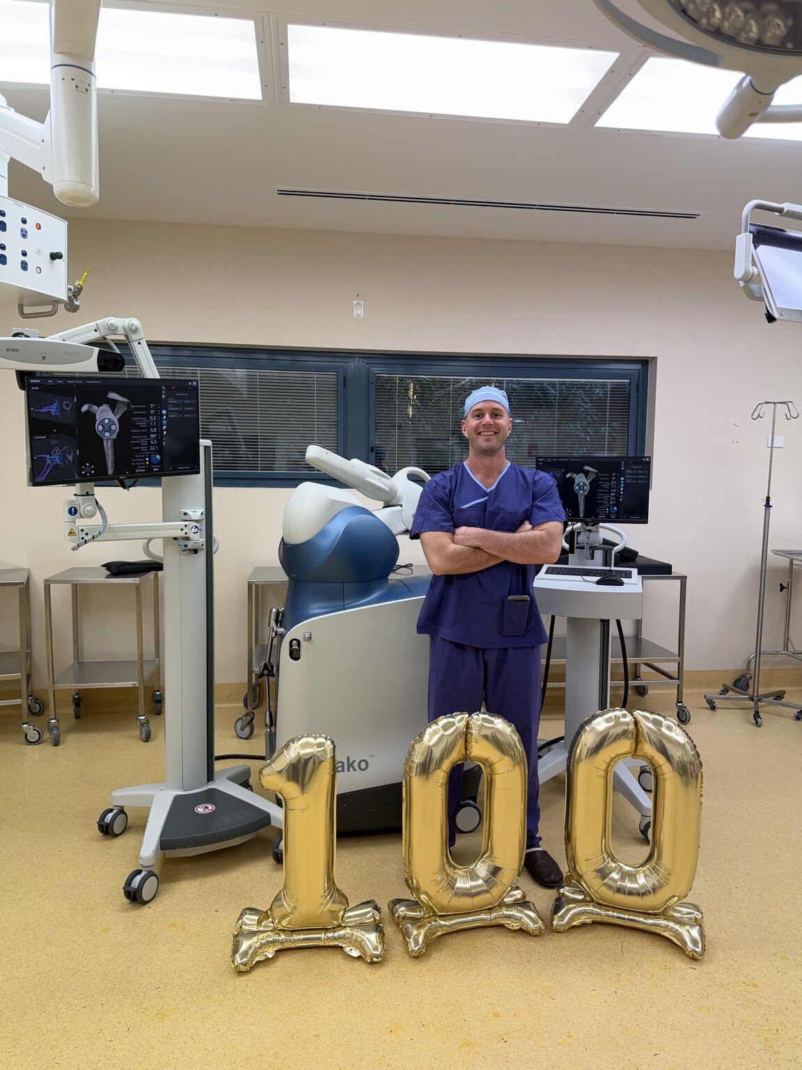 Dr Joe Coory in theatre beside the Mako robotic arm, with gold balloons reading 100, marking his hundredth Mako robotic reverse total shoulder replacement